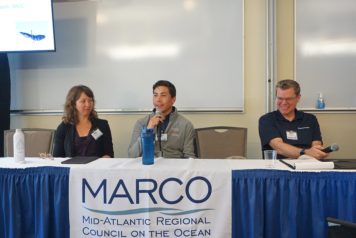 Three panelists speak behind a table covered with a MARCO banner. 